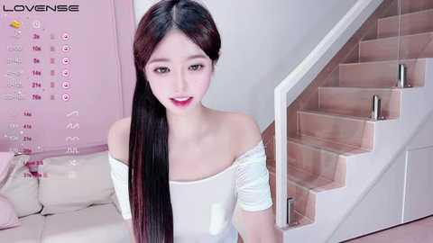 Media: Video of an Asian woman with long black hair, fair skin, and red lipstick, wearing a white off-shoulder dress, standing in a modern, minimalist room with a white staircase and pink accents.