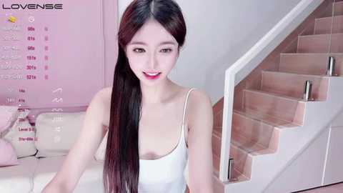 Media: A video of an Asian woman with long, straight black hair, fair skin, and small breasts, wearing a white tank top, sitting on a staircase in a pastel pink room.