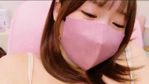 Media: Video of a young Asian woman with long brown hair, wearing a pink face mask and a light pink top, seated in a spa-like environment.