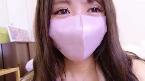 Media: Video of an East Asian woman with long, straight brown hair, wearing a light pink surgical mask, revealing her large, almond-shaped eyes. She is indoors with a wooden wall and a green lamp in the background.