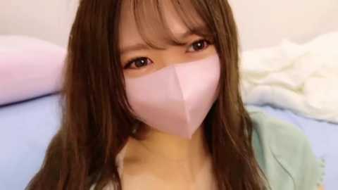 Media: Video of a young Asian woman with long brown hair and fair skin, wearing a pink face mask, lying on a bed with light blue and white sheets, creating a soft, serene atmosphere.