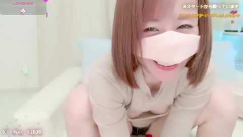 Media: A young East Asian woman with light skin and short brown hair, wearing a pink face mask, is smiling, sitting on a white bed. The room is brightly lit with soft pastel colors.