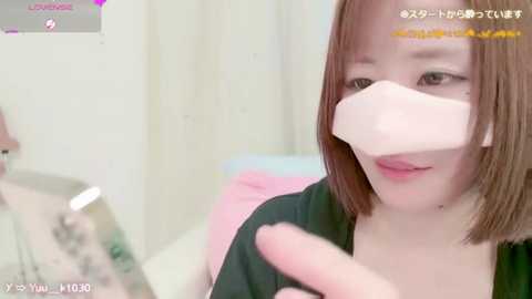 Media: Video of an Asian woman with straight, shoulder-length brown hair, wearing a face mask, black top, and pink blouse, in a brightly lit room, with text overlays in Japanese.