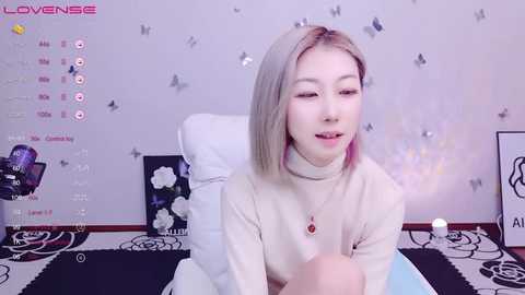 Media: A video of a young Asian woman with straight, shoulder-length platinum blonde hair, wearing a cream-colored turtleneck sweater. She sits on a white chair in a room adorned with butterfly stickers and a black-and-white floral rug.