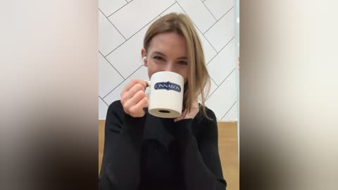 Media: Video of a young woman with light brown hair, wearing a black sweater, holding a white coffee cup with a blue \"Lanaca\" logo, against a white, geometric-patterned wall.