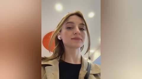 Media: Video of a young Caucasian woman with fair skin, light brown hair, and a neutral expression. She wears a beige trench coat over a black sweater, standing indoors with a blurred background of orange and white abstract art.