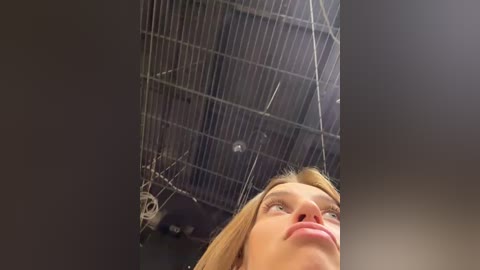 Media: A video captures a low-angle view of a woman with light skin and blonde hair, looking up at a metallic, grid-like ceiling. The background shows industrial elements, wires, and pipes.