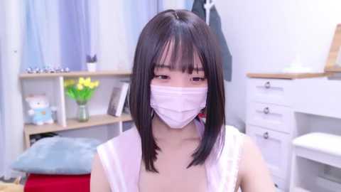 Media: Video of a young Asian woman with straight black hair, wearing a white face mask, white top, in a brightly lit, minimalist bedroom with white furniture, yellow flowers, and a small teddy bear.