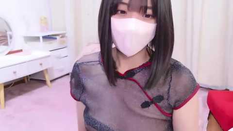 Media: Video of a young Asian woman with shoulder-length black hair, wearing a white mask, a sheer black top with red trim, and seated in a pink room with white furniture and a desk.