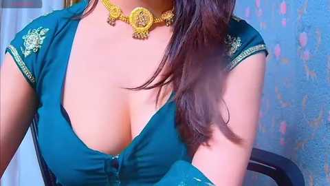 Media: Video of a woman with fair skin, wearing a low-cut green blouse revealing ample cleavage, adorned with a golden necklace and earrings, seated indoors against a blue floral-patterned wall.