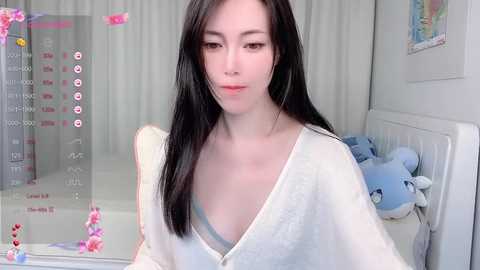 Media: A video of a young woman with long, straight black hair, fair skin, and a slender physique, wearing a white robe, in a cozy, pastel-themed room.