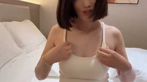 Media: Video of an East Asian woman with shoulder-length black hair, fair skin, wearing a white tank top, adjusting straps. She stands in a modern, beige bedroom with a bed and framed artwork on the wall.