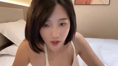 Media: Video of an Asian woman with short black hair, wearing a white top, lying on a bed with white sheets and beige headboard. Background features a modern, minimalist room with a framed abstract painting.