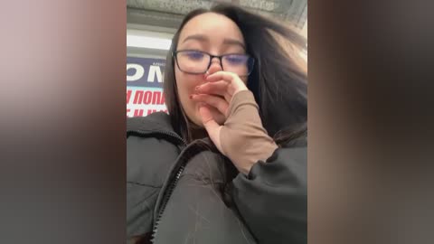 Media: Video of a young woman with long dark hair, glasses, and a black jacket, covering her mouth with her right hand, standing indoors near a sign with red and blue text.