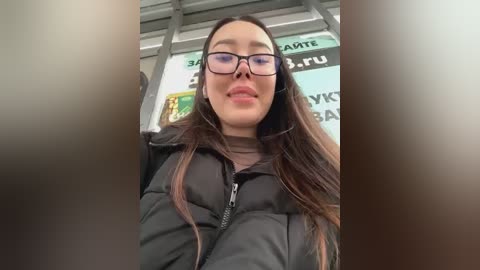 Media: A video of a smiling, fair-skinned young woman with long, straight dark hair, wearing black-framed glasses and a black puffy jacket, seated in a public bus. The background shows a bus window with partially visible advertisements.