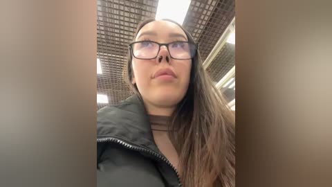 Media: Video of a young woman with long, straight brown hair, wearing glasses, a black puffer jacket, and a beige shirt, looking up with a neutral expression in an indoor setting with tiled ceiling.