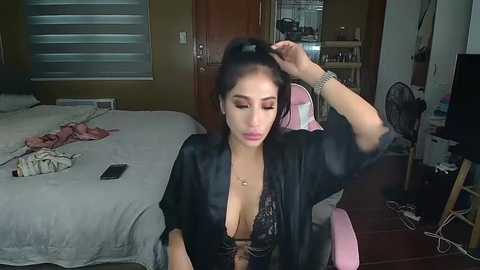 Media: Video of a young woman with light skin, long dark hair, and large breasts, wearing a black lace bra and robe, sitting in a pink gaming chair in a messy bedroom with a bed, fan, and door.