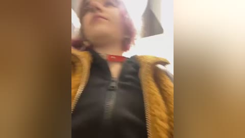 Media: Video of a young woman with short, pink hair, wearing a mustard-yellow jacket over a black shirt with a red collar, standing in a dimly lit room with a white ceiling and a fan.