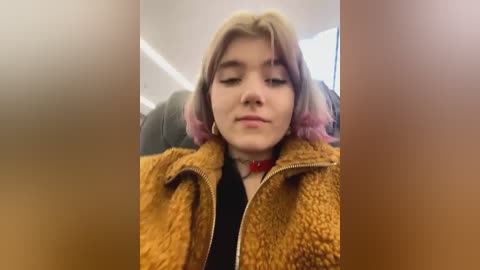 Media: Video of a young woman with shoulder-length, light brown hair, wearing a fuzzy brown jacket and a red choker, seated in a plane with a blurred background.