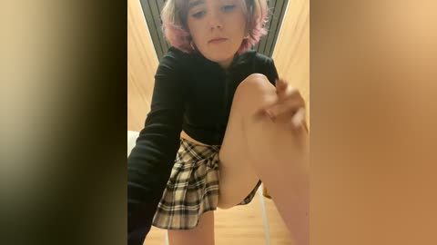 Media: Video of a young person with short, pink hair, wearing a black crop top and plaid skirt, lifting their leg in a wooden hallway, creating a blurred background.