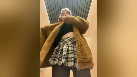 Media: Video of a young woman with platinum blonde hair, wearing a mustard-yellow fuzzy coat, black crop top, and a short black-and-white plaid skirt, standing in a hallway with beige walls and a light ceiling.