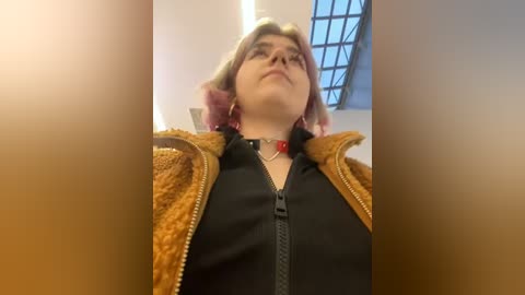 Media: Video of a person with short, pink hair, wearing glasses, a black top, and a brown fur-trimmed jacket, looking upward. Background features a grid window and beige walls.