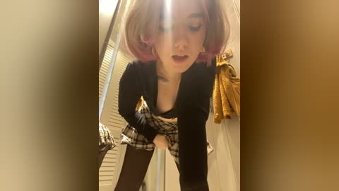 Media: A video of a young woman with shoulder-length, pastel pink hair, wearing a black shirt and a plaid skirt, leaning forward, capturing a candid, slightly blurry moment in a dimly lit room.