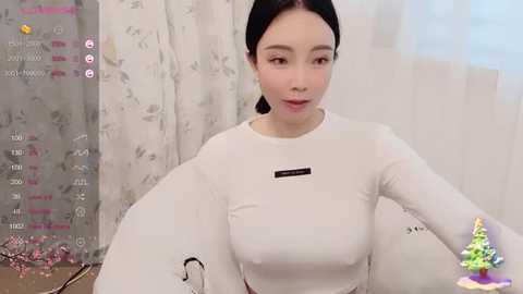 Media: Video of an East Asian woman with fair skin, dark hair, and light makeup, wearing a snug, white, long-sleeved crop top. She sits in a room with patterned curtains and a small Christmas tree in the background.