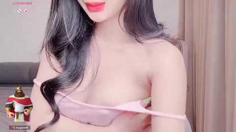 Media: Video of a young woman with long, wavy black hair, wearing a light pink off-shoulder top, red lipstick, and a subtle necklace, in a softly lit bedroom.