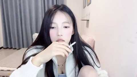 Media: A video of an East Asian woman with long black hair, wearing a white shirt, sitting on a bed, holding a syringe to her mouth, in a modern, minimalistic room with gray curtains.