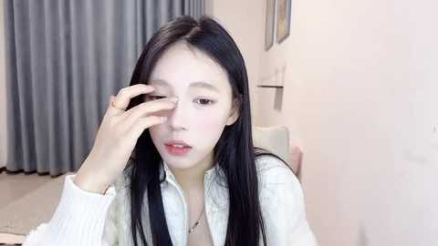 Media: Video of an East Asian woman with long black hair, wearing a white shirt, adjusting her glasses. Background features beige walls, a gray curtain, and a framed picture.