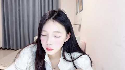 Media: A video of a young Asian woman with long black hair, fair skin, and delicate features, wearing a white blouse, sitting in a bright room with gray curtains and a white wall.