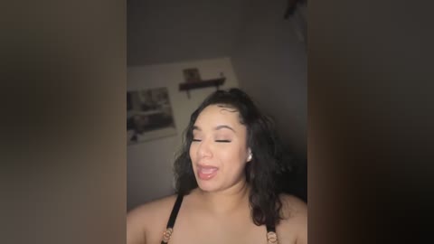 Media: Video of a young woman with curly black hair and light skin, wearing a black bra, sticking out her tongue, in a dimly lit room with a shelf and framed photos in the background.