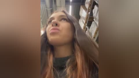 Media: Video of a young woman with light brown skin and long, straight, auburn hair, wearing a black top, posing in a dimly lit, cluttered room with metal shelves and cardboard boxes in the background.