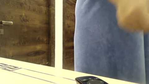 Media: Video of a person in a light blue shirt standing in front of a wooden door with a metal handle. The person's arm is visible, holding a black smartphone. The background features a rustic wooden wall.