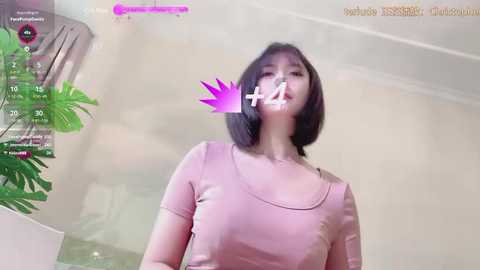 Media: Video of an East Asian woman with short black hair and a pink top, vaping indoors with a pink overlay. Background features a potted plant and a white wall.