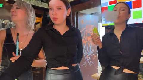 Media: Video of three women in a bar, all wearing black outfits; one with a short, blonde bob, one with brown hair tied back, and one with dark hair. Background features colorful abstract art and a lively bar setting.