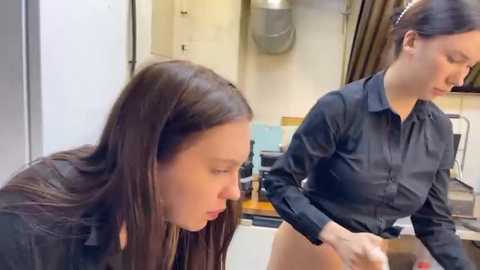 Media: Video of two young women in a kitchen, one with long brown hair and black top, the other with dark hair tied back, wearing a black shirt, both looking down.