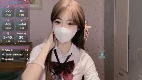 Media: Video of a young Asian woman in a white shirt with a red plaid bow tie, wearing a white mask, adjusting her hair, in a dimly lit room with a calendar and a green light on the wall.