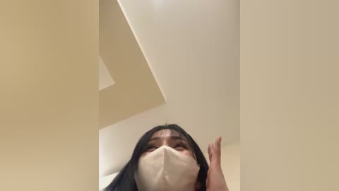 Media: Video of a woman with long black hair and light skin, wearing a white face mask, standing indoors with beige walls and a white ceiling.