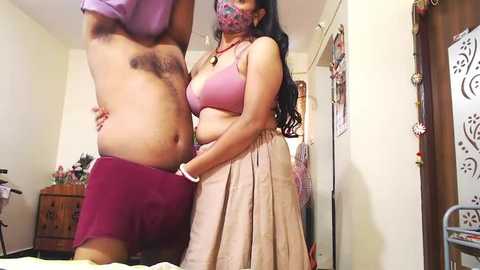 Media: Video of a woman with long black hair, wearing a pink bra and beige skirt, standing next to a man with dark hair, in a bedroom.