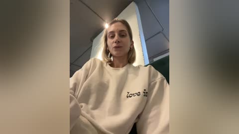 Media: Video of a young woman with blonde hair, wearing a white sweatshirt with Japanese characters, seated in an airplane with overhead compartments and green seats visible.