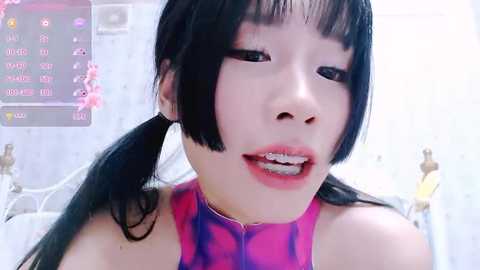 Media: Video of an East Asian woman with long black hair, wearing a pink and purple tie-dye top, showing braces. Background features a pink floral wall and a digital camera screen.