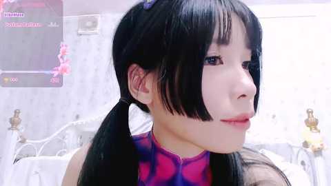 Media: A video of an East Asian woman with long, straight black hair styled in pigtails, wearing a purple and pink cheongsam dress, indoors. The background features a white bed with gold accents.