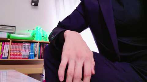 Media: Video of a person in a dark suit sitting on a bed, hand on their knee, in a room with green neon lights, white curtains, and a bookshelf filled with colorful books.