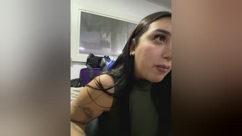 Media: A video of a young woman with olive skin and long black hair, wearing a black turtleneck, looking contemplative. Background shows a cluttered room with purple luggage and a framed artwork.