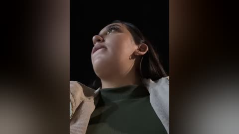 Media: A close-up video of a woman with fair skin, dark hair, and green eyes, wearing a green top and beige jacket, gazing upward against a dark background.