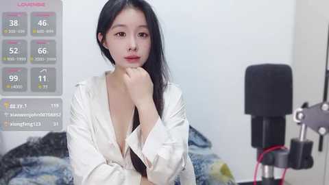 Media: Video of an East Asian woman with long black hair, wearing a white robe, resting her chin on her hand. Background shows a bed with a gray blanket and a microphone stand.