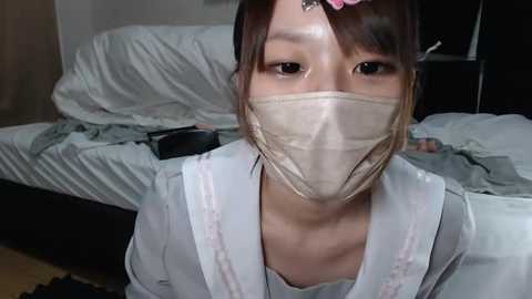 Media: Video of an Asian woman with light skin, wearing a beige surgical mask, pink hair clip, and a white blouse, in a messy bedroom with white sheets and a black dresser.