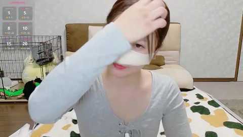 Media: A video of an Asian woman with light skin, brown hair tied back, wearing a light gray cardigan, covering her face with a white cloth. She sits on a bed with a green and brown patterned blanket in a room with a wooden floor and a beige headboard.